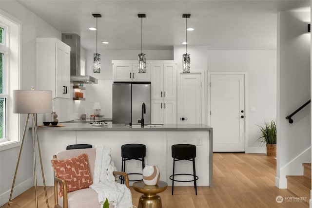 kitchen with pendant lighting, wall chimney range hood, stainless steel fridge, backsplash, and white cabinetry