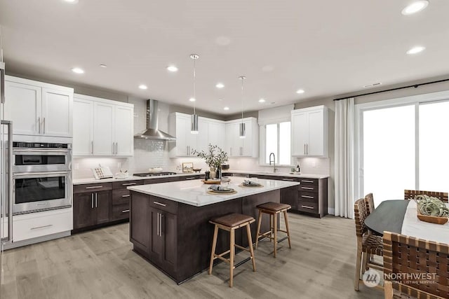 kitchen with stainless steel appliances, light wood-type flooring, a kitchen island, wall chimney range hood, and a breakfast bar