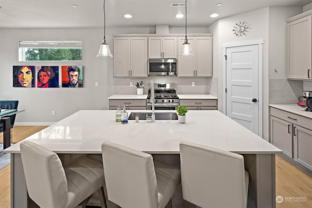 kitchen featuring decorative light fixtures, stainless steel appliances, an island with sink, light wood-type flooring, and gray cabinetry