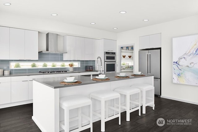 kitchen with stainless steel appliances, a kitchen island with sink, dark wood-type flooring, wall chimney range hood, and white cabinets