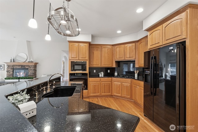 kitchen featuring dark stone counters, a sink, a stone fireplace, light wood-type flooring, and black appliances