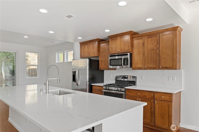 kitchen with a sink, stainless steel appliances, a kitchen island with sink, and brown cabinetry