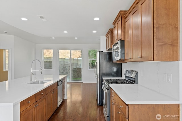 kitchen featuring a sink, brown cabinetry, dark wood-style floors, stainless steel appliances, and a kitchen island with sink