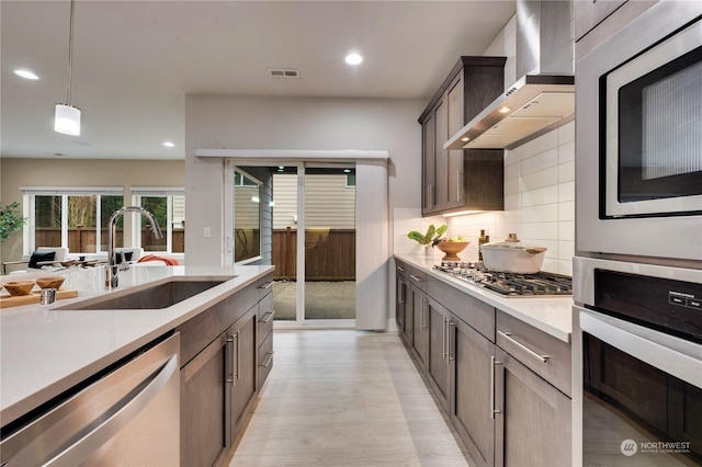 kitchen featuring wall chimney exhaust hood, hanging light fixtures, stainless steel appliances, tasteful backsplash, and sink