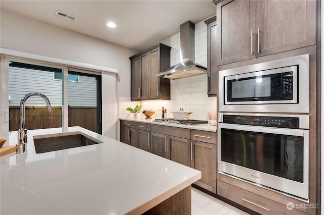 kitchen with appliances with stainless steel finishes, dark brown cabinets, wall chimney range hood, and sink