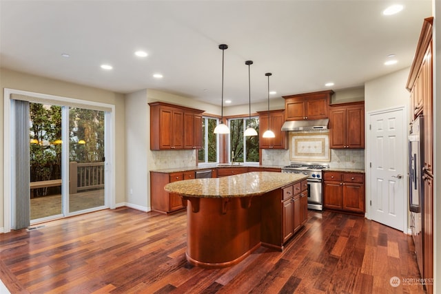 kitchen with appliances with stainless steel finishes, dark wood-type flooring, a kitchen island, pendant lighting, and light stone counters