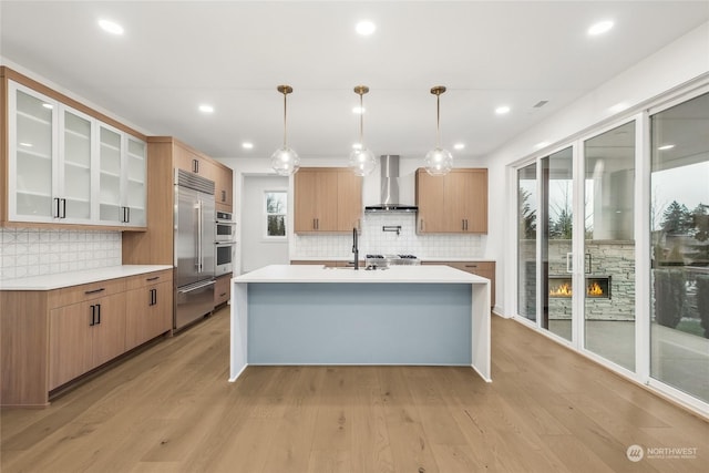 kitchen featuring light hardwood / wood-style flooring, hanging light fixtures, wall chimney range hood, appliances with stainless steel finishes, and a stone fireplace