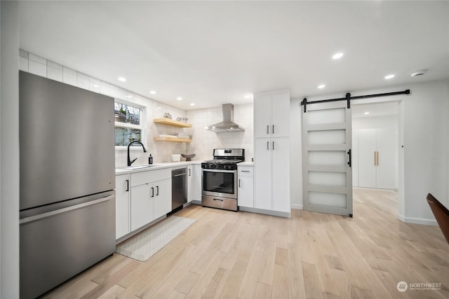 kitchen featuring a barn door, appliances with stainless steel finishes, wall chimney range hood, white cabinets, and sink