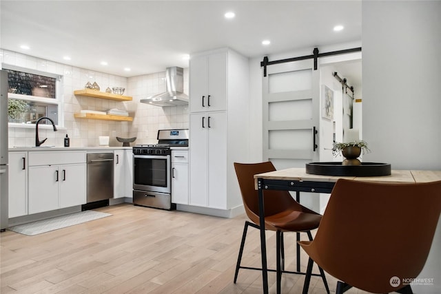 kitchen featuring white cabinetry, a barn door, stainless steel appliances, wall chimney range hood, and light hardwood / wood-style flooring