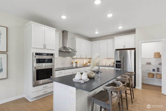 kitchen featuring an island with sink, wall chimney range hood, white cabinets, appliances with stainless steel finishes, and a kitchen breakfast bar