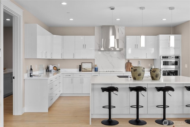 kitchen featuring a breakfast bar, white cabinets, wall chimney exhaust hood, and stainless steel double oven