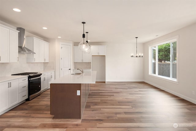 kitchen featuring wall chimney range hood, white cabinets, pendant lighting, stainless steel gas stove, and an island with sink