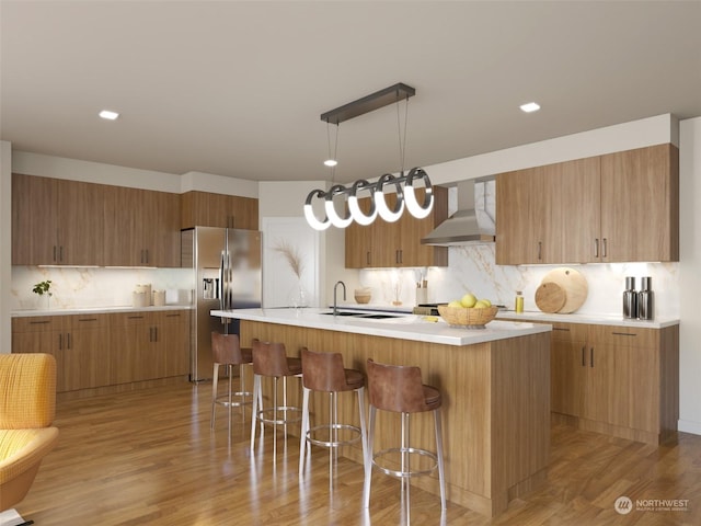 kitchen featuring light hardwood / wood-style flooring, wall chimney range hood, stainless steel fridge, a kitchen island with sink, and backsplash