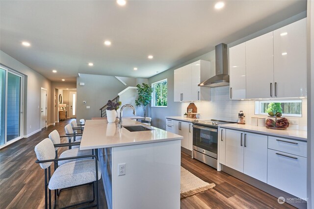 kitchen featuring a sink, white cabinets, stainless steel range with electric cooktop, wall chimney exhaust hood, and an island with sink