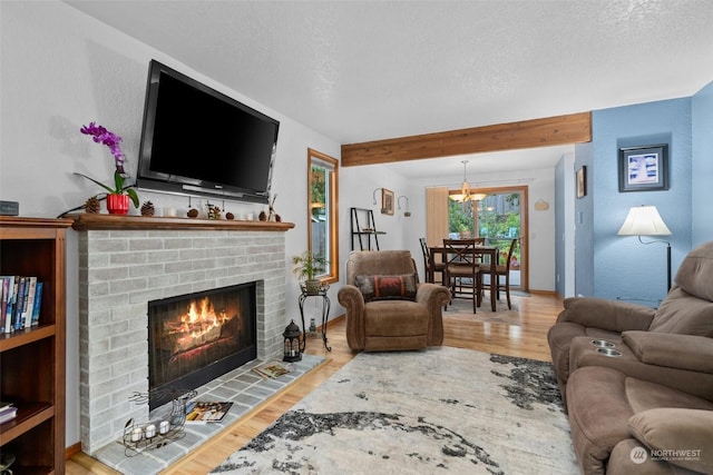 living room with a brick fireplace, a textured ceiling, a chandelier, light hardwood / wood-style flooring, and beamed ceiling