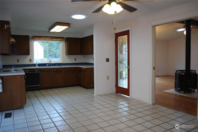 kitchen with sink, light tile patterned floors, ceiling fan, black dishwasher, and a wood stove