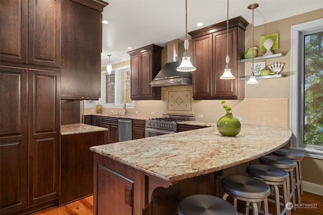 kitchen featuring appliances with stainless steel finishes, hanging light fixtures, dark brown cabinets, wall chimney exhaust hood, and a kitchen bar