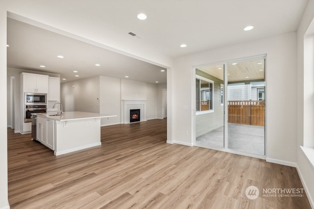 kitchen featuring white cabinetry, an island with sink, oven, light wood-type flooring, and built in microwave