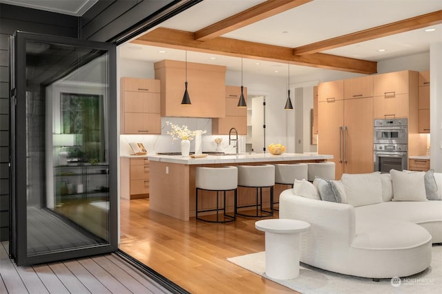 kitchen with light brown cabinets, light hardwood / wood-style flooring, hanging light fixtures, a kitchen island with sink, and stainless steel double oven