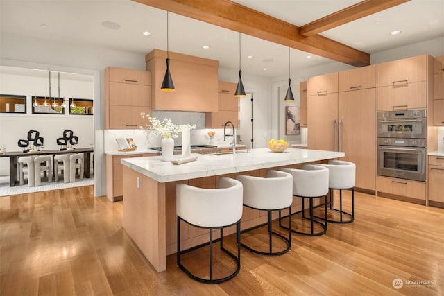 kitchen with a kitchen island with sink, stainless steel double oven, pendant lighting, and light brown cabinets