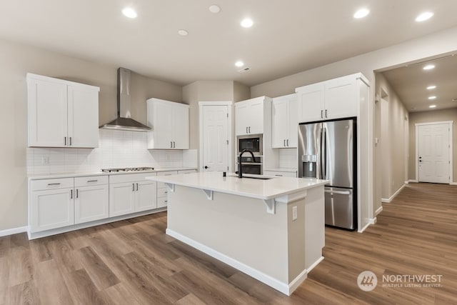 kitchen featuring stainless steel refrigerator with ice dispenser, white cabinetry, a kitchen island with sink, and wall chimney range hood