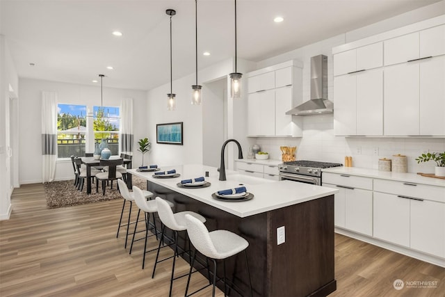 kitchen with high end stove, wall chimney exhaust hood, an island with sink, and white cabinetry