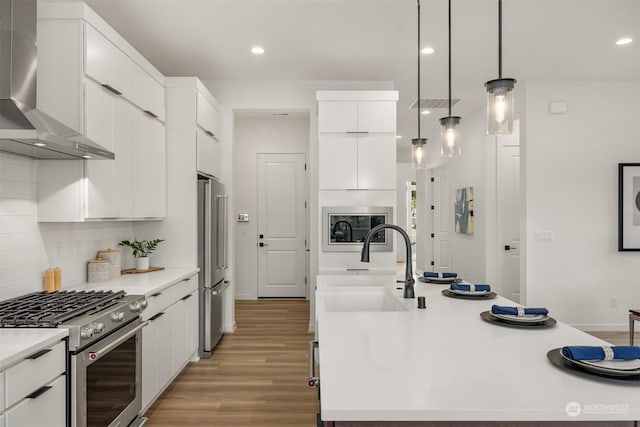 kitchen featuring wall chimney range hood, hanging light fixtures, light wood-type flooring, appliances with stainless steel finishes, and white cabinets