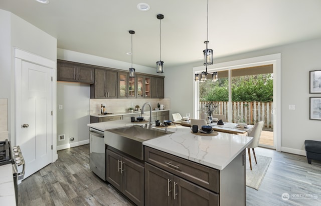 kitchen with light stone counters, decorative light fixtures, dark brown cabinets, appliances with stainless steel finishes, and a kitchen island with sink