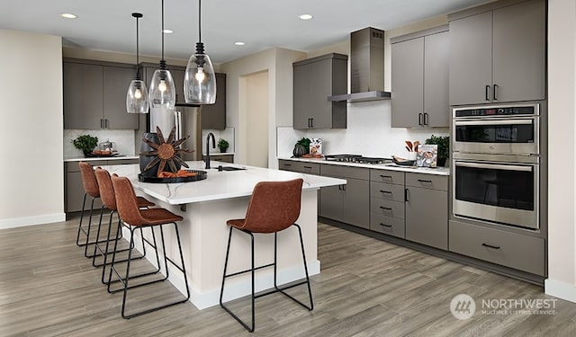 kitchen with wall chimney range hood, appliances with stainless steel finishes, gray cabinets, and a center island with sink