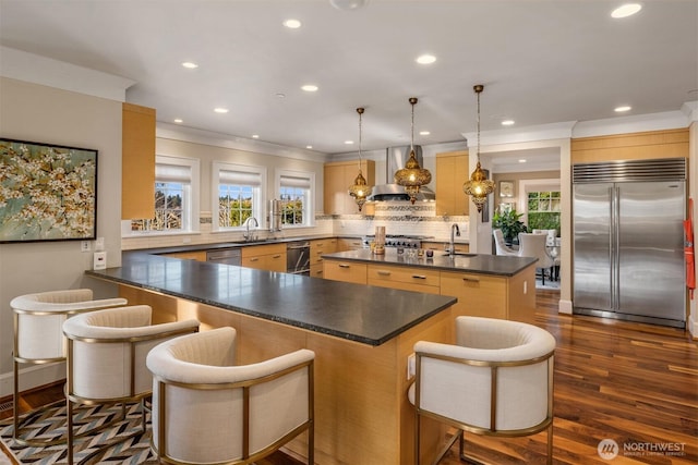 kitchen featuring dark countertops, backsplash, wall chimney range hood, a center island with sink, and appliances with stainless steel finishes