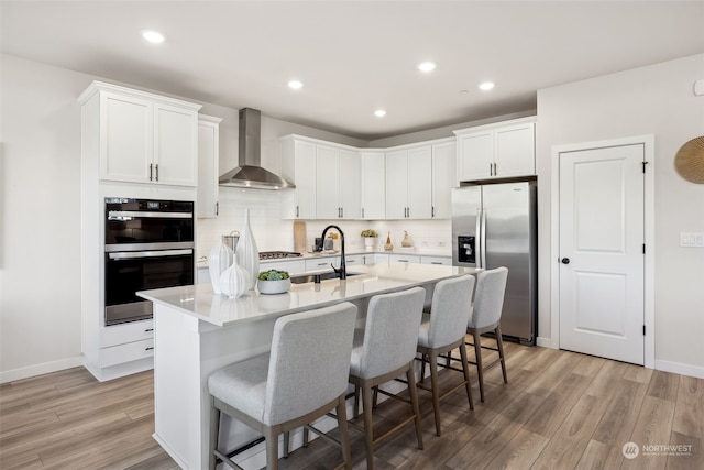 kitchen featuring wall chimney exhaust hood, sink, stainless steel fridge with ice dispenser, an island with sink, and white cabinets