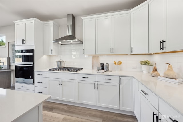 kitchen featuring backsplash, double wall oven, stainless steel gas cooktop, white cabinets, and wall chimney exhaust hood