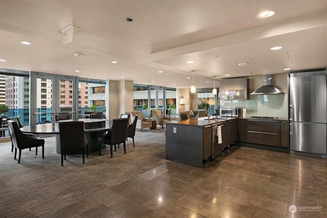 kitchen with wall chimney exhaust hood, stainless steel appliances, tasteful backsplash, sink, and hanging light fixtures