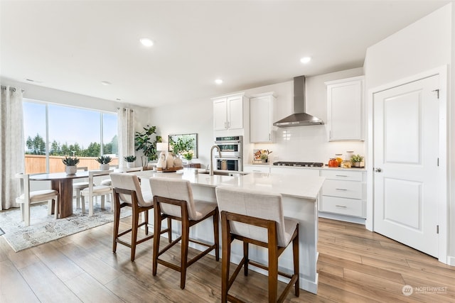 kitchen with white cabinetry, wall chimney exhaust hood, light hardwood / wood-style floors, and an island with sink