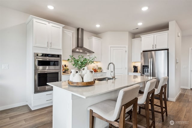 kitchen with sink, white cabinetry, appliances with stainless steel finishes, a kitchen island with sink, and wall chimney range hood