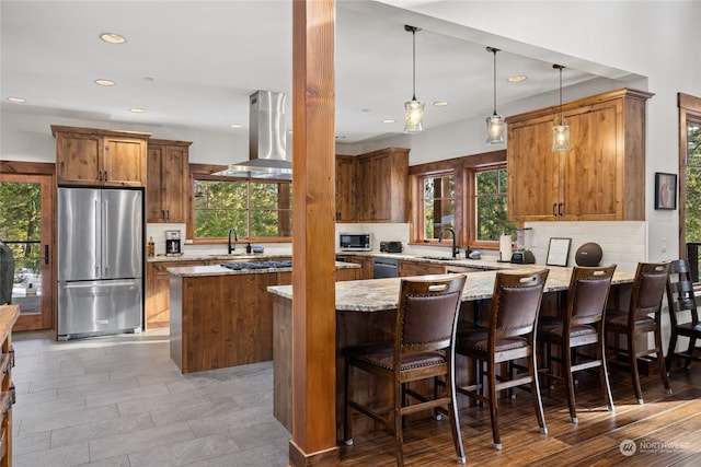 kitchen featuring hanging light fixtures, stainless steel appliances, island range hood, light stone countertops, and a kitchen island