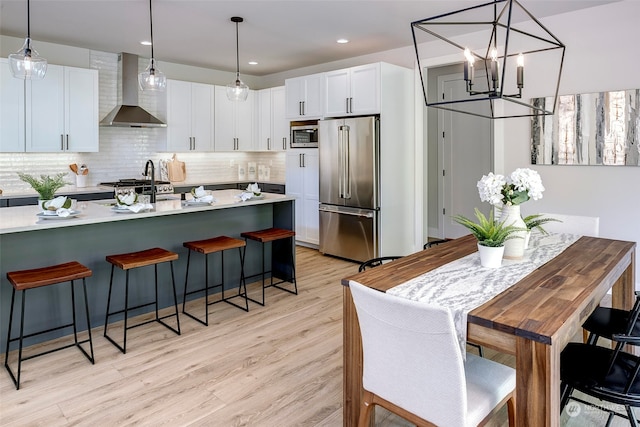 kitchen with appliances with stainless steel finishes, white cabinets, pendant lighting, wall chimney exhaust hood, and light hardwood / wood-style flooring