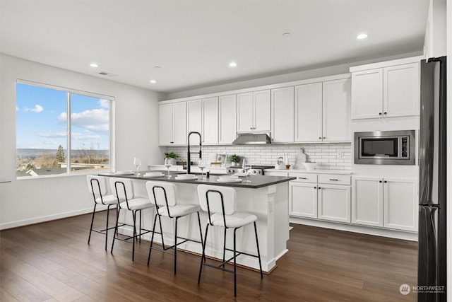 kitchen featuring stainless steel microwave, a kitchen bar, white cabinetry, a kitchen island with sink, and black fridge