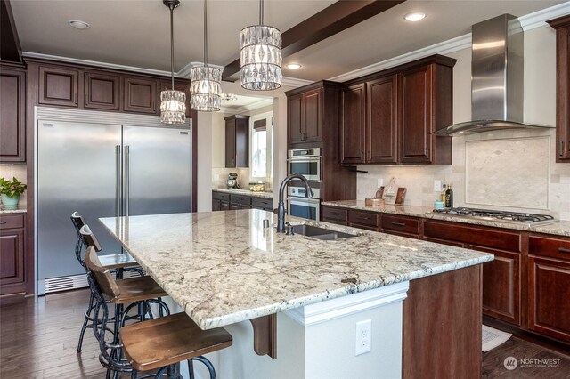 kitchen with light stone counters, an island with sink, a sink, appliances with stainless steel finishes, and wall chimney range hood