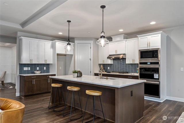 kitchen with an island with sink, stainless steel appliances, decorative light fixtures, dark wood-type flooring, and white cabinets