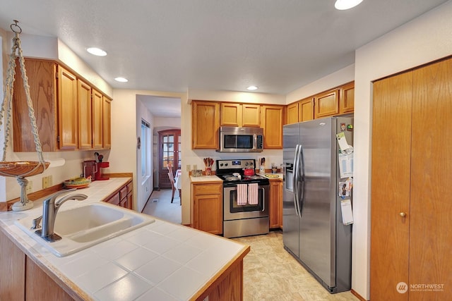 kitchen featuring tile counters, brown cabinetry, a peninsula, stainless steel appliances, and a sink