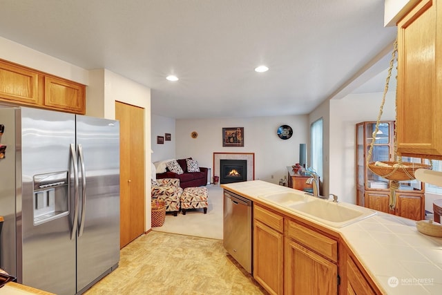 kitchen featuring tile counters, appliances with stainless steel finishes, brown cabinetry, a sink, and a lit fireplace