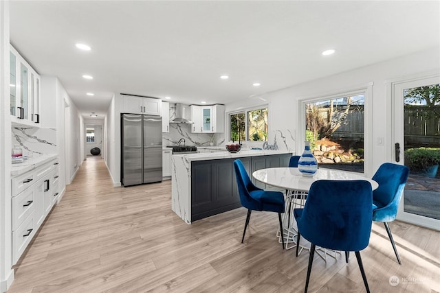 kitchen featuring wall chimney range hood, light hardwood / wood-style flooring, stainless steel refrigerator, white cabinets, and decorative backsplash