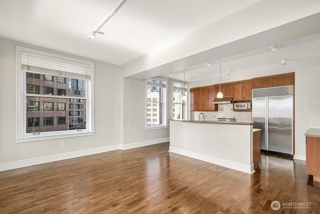 kitchen with under cabinet range hood, dark wood-style flooring, appliances with stainless steel finishes, decorative backsplash, and brown cabinetry