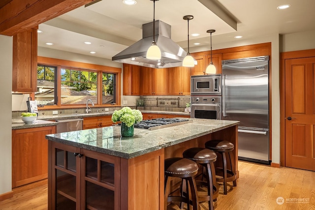 kitchen with pendant lighting, light stone countertops, a kitchen island, island range hood, and built in appliances