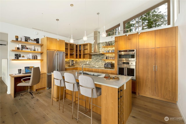 kitchen with open shelves, appliances with stainless steel finishes, brown cabinetry, light wood-type flooring, and wall chimney exhaust hood