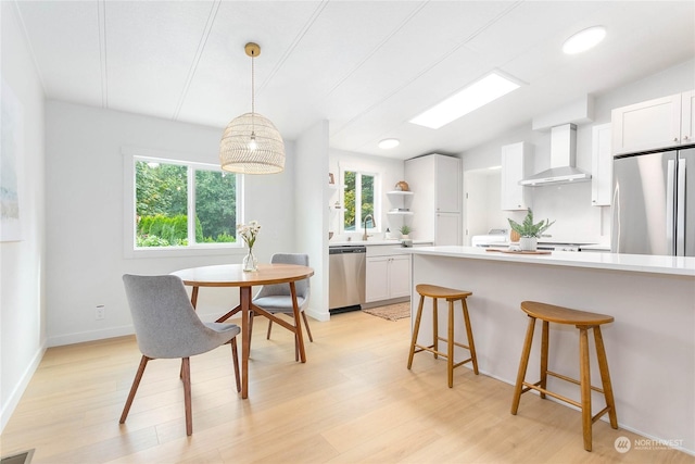 kitchen with white cabinets, appliances with stainless steel finishes, wall chimney exhaust hood, hanging light fixtures, and light wood-type flooring