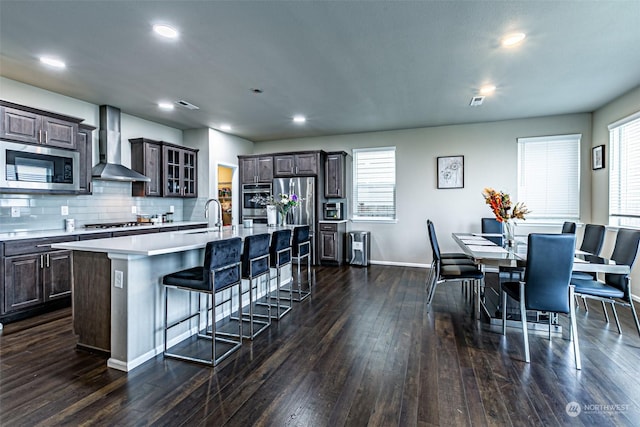 kitchen with wall chimney exhaust hood, a center island with sink, dark brown cabinets, and appliances with stainless steel finishes