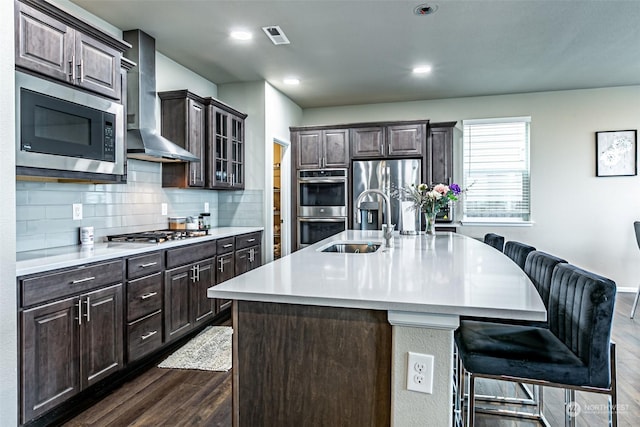 kitchen featuring dark wood-type flooring, wall chimney range hood, stainless steel appliances, an island with sink, and a kitchen breakfast bar