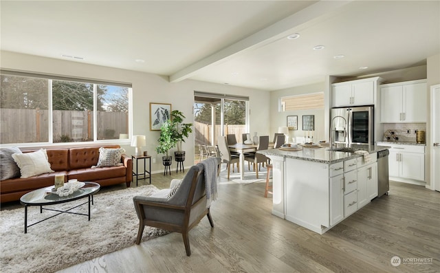 kitchen with white cabinetry, appliances with stainless steel finishes, beamed ceiling, light stone countertops, and a kitchen island with sink